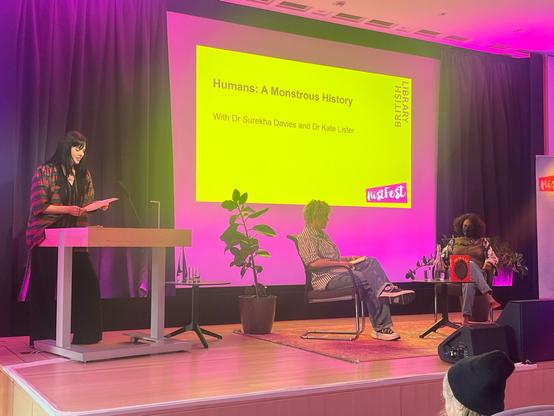 Stage with giant, bright yellow screen with pink border. Three women on stage: at left, one at the podium; in the centre, two seated in front of screen, around a small table with a bottle of water, two glasses, and a bright red book with dark oval circle on the cover sits on the table.