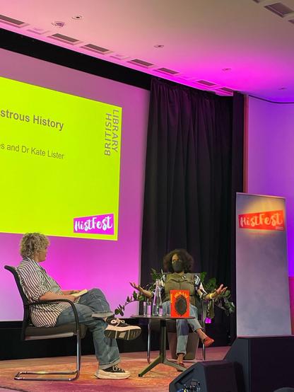 Stage with giant, bright yellow screen with pink border. Two women seated in front of screen, around a small table with a bottle of water, two glasses, and a bright red book with dark oval circle on the cover sits on the table. To the right is a pale blue banner with pink logo: HistFest.