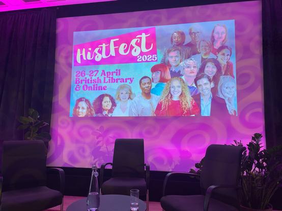 Stage with giant screen showing psychedelic blue and pink HistFest poster with pink logo with white writing. The bottom half along the diagonal (screen divided bottom left to topi right) is an array of authors’ headshots. The screen is behind a stage with three chairs around a table.