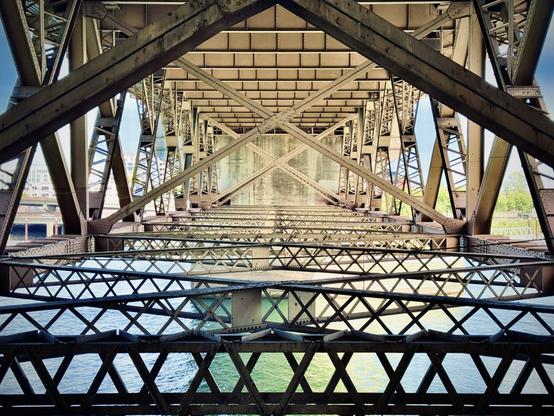Layers of crisscrossing beams meet at perpendicular joints under a large, concrete bridge over shiny emerald waters on a sunlit day.