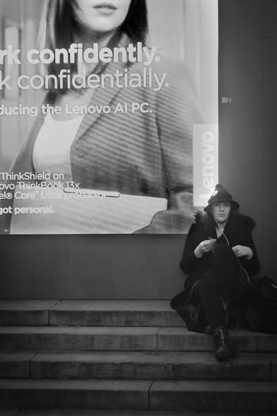 On the right of the picture a woman dressed all in black with a hat shaped like those worn by a witch is sitting facing the viewer on some steps and knitting something, a spell, perhaps. She has a large black tote bag at her left side and the cord of her white earbuds reaches down inside it. On the dark grey wall behind her is a video screen advertising Lenovo PCs.