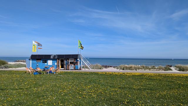 Blick vom Deich auf die Surfschule Heidkate, das Gebäude ist mit Holz verkleidet und blau gestrichen. Es wehen Fahnen an der Surfschule. Davor stehen Liegestühle. 
Im Vordergrund ist grünes Gras mit gelben Löwenzahn. 
Im Hintergrund die blaue Ostsee, der Himmel ist blau mit einigen Schleierwolken.