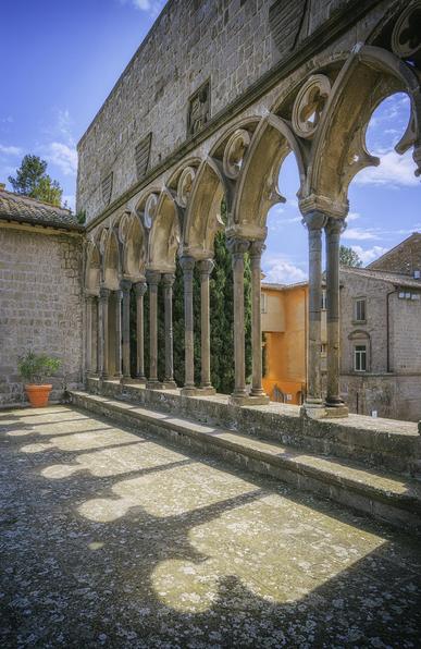 The arches of the loggia of the papal palace in Viterbo Italy cast shadows on the patio below. There are 6 arches receding away from the viewer, with a view of some of the city's buildings through the openings. Blue sky with white puffy clouds can be seen.
