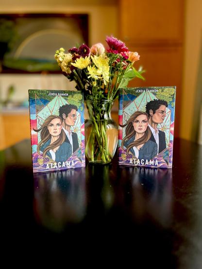 A vase of mixed flowers on a black table, with the novel Atacama by Jendia Gammon on either side; the paperback is on the left and the hardcover is on the right.