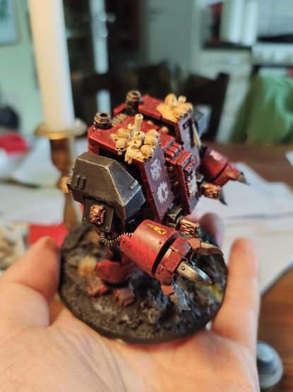 It's a Warhammer 40k Dreadnought, basically a gorilla shaped walking coffin with various dangerous weapons attache, photographed from a 3/4 top down right side angle. It's painted a dark red with black, and has skulls and candles on its shoulders.