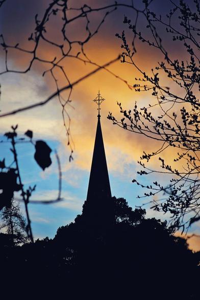 A silhouette of a church steeple topped with a cross stands against a vibrant sky with hues of orange, yellow, and blue, framed by dark, bare tree branches in the foreground.