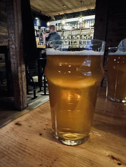A glass of Malduguns Clipa APA beer sits on a wooden table in the cozy atmosphere of the pub “Alus muiža” in Riga. The beer glows with a golden hue under soft lighting, with a frothy head partially clinging to the glass. Other patrons are visible in the dimly lit background, adding to the lively yet relaxed ambiance.