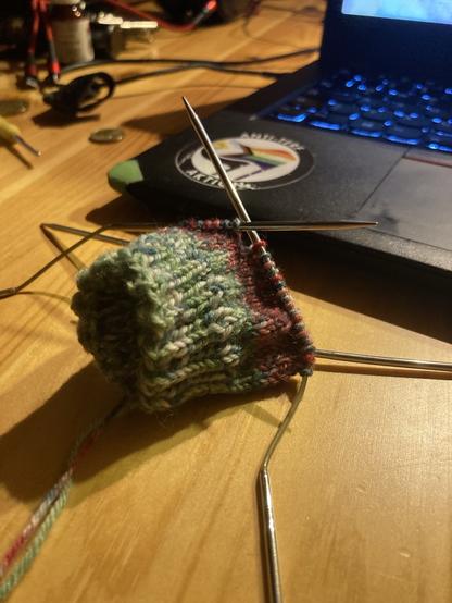 Eine angefangene Socke aus grün-weiß-roter Wolle.