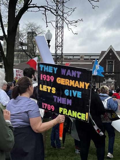 A woman holding a colorful protest sign that says, "They want 1939 Germany, let's give them 1789 France!" The scene is set outdoors with cloudy skies.