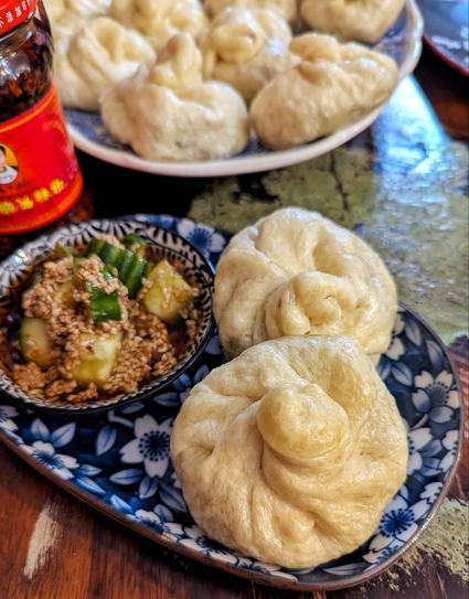 Holztisch, darauf ein ovaler Teller mit blauem Kirschblütenmotiv. auf dem Teller 2 große asiatische, herzhaft gefüllte Dampfnudeln, daneben ein Schälchen mit asiatischem Gurkensalat. Die Gurkenstücke schwimmen in einem braunen Dressing mit Sesamsaat. Dahinter verschwommen ein Glas mit Crispy Chili Oil und eine große Porzellanplatte mit weiteren Dumplings.