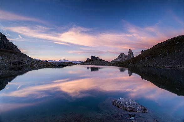 Photo d'un paysage de montagne au lever du soleil.
Un caillou baigne dans un lac, on devine à la profondeur que la berge est toute proche. 
Au bout du lac se trouve un refuge sur un petit promontoire encadré de part et d'autre par deux collines, juste à sa droite se dresse une montagne en forme de canine. Juste à gauche du refuge, on distingue très loin à l'horizon des montagnes dont certaines sont enneigées.
Le ciel est bleu, traversé de fins nuages rose pâle qui se reflètent dans le lac.
-
Photo of a mountain landscape at sunrise.
A pebble lies in a lake, and you can tell by its depth that the shore is nearby. 
At the end of the lake stands a refuge on a small promontory framed on either side by two hills, with a canine-shaped mountain just to its right. Just to the left of the hut, you can make out mountains in the distance, some of them snow-capped.
The sky is blue, with fine pale pink clouds reflected in the lake.