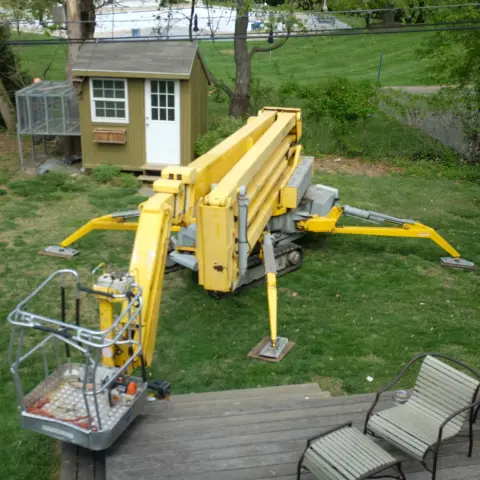 A yellow insect-like tracked crane with corner stabilizers, nesting in a yard