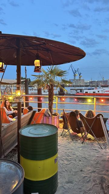 Ein Sonnenschirm, eine Metalltonne, eine Reihe von Liegestühlen mit Sand am Boden. Im Hintergrund das Panorama des Hamburger Hafens am Abend. Bar Strand Pauli – Hamburg