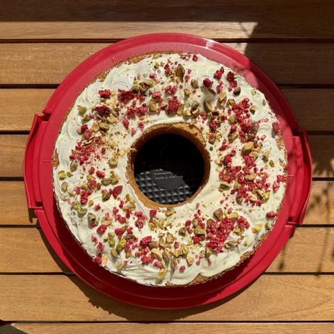 A round cake with a central hole, topped with cream frosting, crushed raspberries, and chopped pistachios, resting on a red cake carrier. The background features wooden surfaces.
