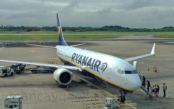Side view of a white and blue, twin engined jet airliner parked facing to the right.
The plane is mostly white, with a blue belly and tail, with yellow piping on the body, and dark blue "RyanAir" billboard-style titles on the upper forward fuselage next to a blue, winged Irish harp.
The same harp appears in yellow on the dark blue tail.
A grey electrical generator is plugged into the planes nose below the cockpit, while a built-in set of airstairs is extended from the forward  on the opposite side, with passengers climbing up into the plane.
There is a luggage belt loader parked against an open door in the lower right rear fuselage, with a luggage cart parked nearby, with 2 men in hi-viz jackets loading bags onto the belt.
Grey concrete apron with white and yellow markings fills the lower half of the frame, with grass, a black runway,  and trees under cloudy grey skies at the top.