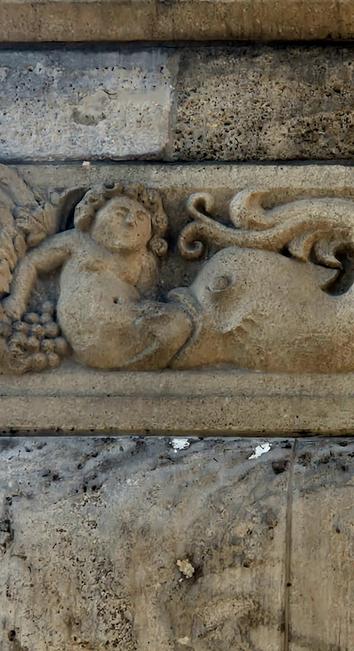Detail of a stone relief on a building façade. It shows a large fish swallowing a reclining human figure from the lower body, with only the upper torso and head still visible. The human seems calm or resigned. Grapes and vine motifs decorate the left side. Above this relief, other sculpted elements include a stylized eagle in an octagonal frame and decorative ornaments.