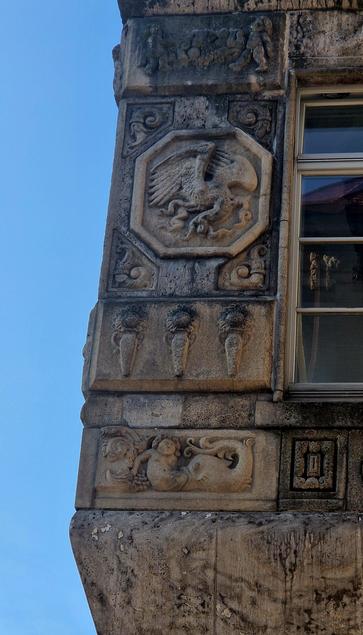 Detail of a stone relief on a building façade. It shows a large fish swallowing a reclining human figure from the lower body, with only the upper torso and head still visible. The human seems calm or resigned. Grapes and vine motifs decorate the left side. Above this relief, other sculpted elements include a stylized eagle in an octagonal frame and decorative ornaments.