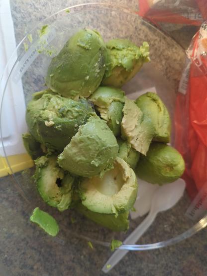 The image shows a close-up of a creamy, greenish-yellow mixture, likely a guacamole or similar dip. The texture appears thick and chunky, with visible pieces of avocado and herbs. The mixture has a smooth, creamy consistency with some lumps, indicating it has been partially mashed. There are flecks of green herbs, possibly cilantro, and small bits of red, which could be onions or tomatoes. The background is indistinct, focusing attention on the mixture itself. The overall appearance suggests a homemade, fresh preparation.
