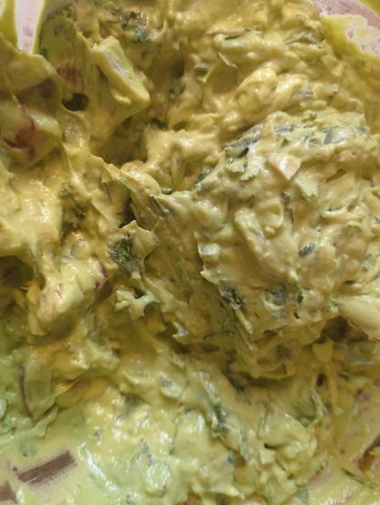 The image shows a clear plastic bowl containing several pieces of avocado. The avocados are green with some light beige flesh visible, indicating they are ripe. The bowl is placed on a granite countertop, and a white plastic spoon is partially visible inside the bowl. A yellow-handled knife is also present, with some avocado residue on it. In the background, there is a red plastic bag, possibly containing bread, and a white cutting board. The avocados appear to have been cut in half and scooped out, with some pieces still having the pit visible. The overall scene suggests that the avocados are being prepared for a recipe.