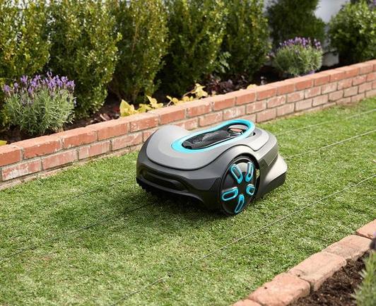 Oblique view of the Gardena Sileno Flex Robotic Lawn Mower from above as it mows a strip of grass bordered by small brick walls. The mower is black with two large wheels in the front - the back wheels underneath the mower are not visible. The mower is a rounded rectangle with curvature around the wheels and on top for a sleek modern look. Turquoise highlights are shown as radial spokes on the wheels. A turquoise oval on top of the mower surrounds a closed panel that covers the controls for the mower. The grass is trimmed short and various bushes are planted on either side of the strip of lawn.