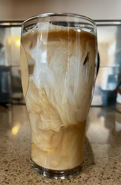 An iced coffee in a glass sits on a counter. The milk and cream is still being mixed in.