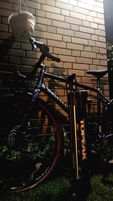 Ein lila-farbenes Gravelbike lehnt an einer Hauswand, davor eine große Luftpumpe. Es ist schon dunkel und eine Außenwandleuchte belichtet die Szenerie von oben.