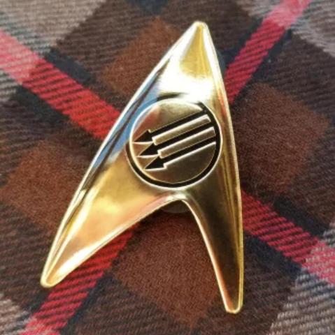 A gold Star Trek Delta badge with a circular emblem in the center displaying three black arrows pointing diagonally downward to the left, representing the anti-fascist symbol. The badge is placed on a brown and red plaid fabric background.