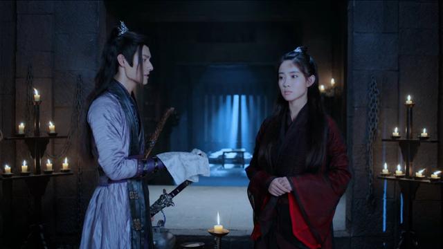 In a Wen dungeon, Jiang Cheng asks Wen Qing a question, and makes an offer. Each wall of the room has a multi-tier stand of lit candles in front of it. Jiang Cheng stands in profile, facing Wen Qing with an outstretched hand coerced in silk handkerchief in which nestles a small object not clearly visible. Wen Qing stands in profile to Jiang Check, staring straight ahead instead of looking at him or the object he holds, hands clasped together at her waist. Blemishes mar the skin on her forehead and throat, possibly smudges of dirt, possibly shadows of bruising.