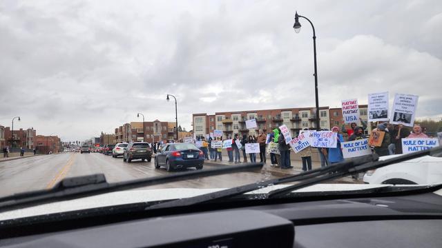 The image shows a protest scene viewed from inside a vehicle. In the foreground, the dashboard and windshield wipers are visible, with raindrops on the windshield. The scene is set on a street with a group of people standing on the sidewalk, holding signs. The protesters are wearing various types of clothing, including jackets and hoodies, suggesting a cool or rainy day. The signs they are holding are not fully legible, but one sign reads "HAND OFF." The background features a multi-story brick apartment building with balconies, and several cars are parked or moving on the street. The sky is overcast, and the overall atmosphere appears to be that of a peaceful demonstration.
