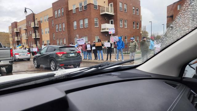 The image depicts a street scene with a group of people standing on the sidewalk, holding signs during a protest. The protest appears to be taking place on a cloudy day, with overcast skies and wet road surfaces, suggesting recent rain. The participants are holding various signs with messages such as "Rule of Law Matters." The signs are predominantly white with blue and red text, and some include images. The protesters are standing in a line, facing the road, with several cars stopped behind them, indicating a traffic stoppage. The background features a row of multi-story buildings with a brick facade, typical of urban residential architecture. The scene is viewed from inside a vehicle, as indicated by the visible dashboard and windshield wipers in the foreground.