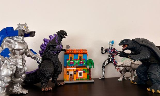 The image shows a collection of toy figures arranged on a dark surface. From left to right: a silver Mechagodzilla with blue accens, a black Shin Godzilla with purple spines, a small colorful LEGO house with flowers and windows, a silver Ultraman Arc with blue accents, a white icy wolf-like creature, and a large Gamera figure. The toys appear to be positioned as if in a standoff, with the house in the middle of the figures.​​​​​​​​​​​​​​​​