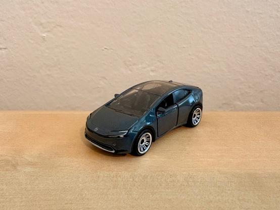 This is a small die-cast toy car model on a wooden surface, a gray Toyota Prius Prime with a hatchback design. The car has black windows and silver wheel rims. The model sits on a light brown wooden table with a beige wall visible in the background.​​​​​​​​​​​​​​​​