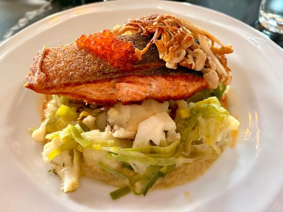 A pan-seared salmon fillet with crispy skin, topped with bright red roe and mushrooms. The fish rests on a bed of braised cabbage and cauliflower in a light broth on a white plate. The salmon is perfectly cooked with moist pink flesh contrasting with the golden-brown exterior.​​​​​​​​​​​​​​​​