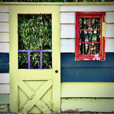 A bright-safety-yellow, glassless wooden door holds a portion of the viewing window's purple-painted pane frames within, as it stands next to a bright red, glassless wooden window, whose housing also holds a portion of its pane frames within, both of which share a wall of wooden slats (from colors white, down to blue, down to safety-yellow), and both of which lead into a grove of bamboo trees beyond their respective thresholds.