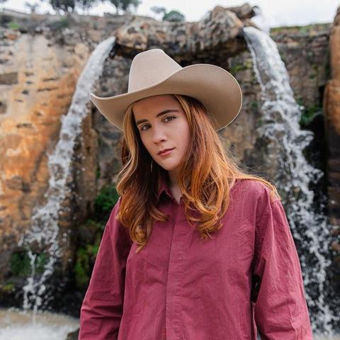 A white woman in a white cowboy hat and loose-fitting red linen shirt looks into the camera. She has long auburn hair. She is framed by a waterfall in the background.