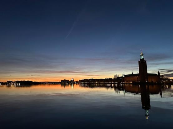 A view over a body of water (Riddarfjärden in Stockholm) westward towards the setting sun. On the right is the City Hall, a large building with a tower, and beyond that, the silhouettes of other buildings, dotted with lights, all the way to the horizon.  In the distance, around the centre, a brightly lit skyscraper stands in front of the glow of the sunset. The water reflects what’s above it.