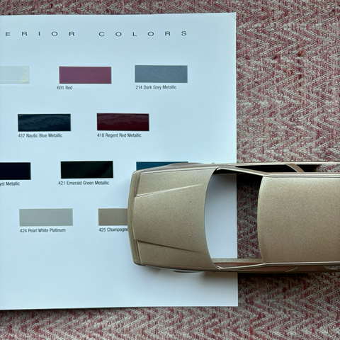 A top-down view of the painted plastic body of a 1:24-scale Tamiya model kit of a Volvo 850 Turbo station wagon, overlaid over the Champagne Metallic color chip on the inside back page of a 1996 Volvo 850 sales brochure, to show the color match with the body.