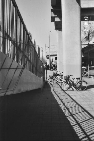 ERA100 E.I 80

- **English:** The image shows a sidewalk next to a tall concrete pillar and a metal fence. Several bicycles are parked near the pillar, casting shadows on the ground. In the background, there are people and vehicles on the street, with an overpass visible above.

- **Chinese (中文):** 这张图片展示了一条人行道，旁边有一个高大的混凝土柱子和一个金属围栏。几辆自行车停在柱子附近，在地面上投下阴影。背景中有街上的人和车辆，上方可见一座高架桥。