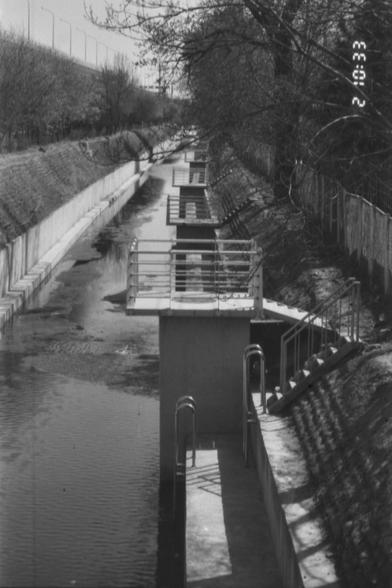 ERA100 E.I 80

- **English:** The image depicts a canal with concrete platforms and railings along its length. Ladders are attached to the platforms, providing access to the water. The canal is bordered by trees and a fence on one side, and a grassy embankment on the other. The image appears in black and white, with a timestamp visible in the upper right corner.

- **Chinese (中文):** 这张图片展示了一条运河，其长度上有混凝土平台和栏杆。平台上安装了梯子，方便进入水面。运河的一侧被树木和围栏包围，另一侧则是草地堤岸。整个图像呈现黑白色调，右上角可见时间戳。