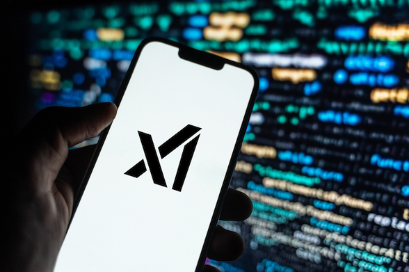 A photo of someone holding a mobile phone with the xAI logo on a white background. Behind the phone is some blurry green white and beige text on a computer screen.