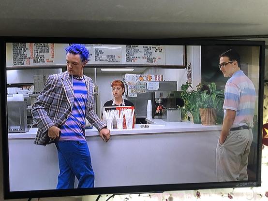 Two punks at a generic 80s fast food joint. The guy on the right has blue hair and is wearing blue pants, a wild plaid jacket, and a pink and blue striped shirt. The pink on the right is in nerd glasses, and is wearing a polo with broad white, pink, and pastel blue stripes with khakis.
