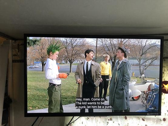 A punk with 12" green hair spikes wearing orange cooking mitts stands before a couple guys in they described outfits, with mopeds in the background. 

"Hey, man. Come on
The kid wants to be a punk, let him be a punk."