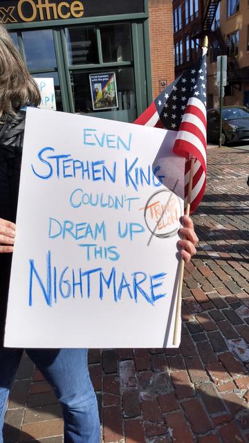 A person holding up a sign and an American flag. The sign reads: "Even Stephen King Couldn't Dream Up This Nightmare"