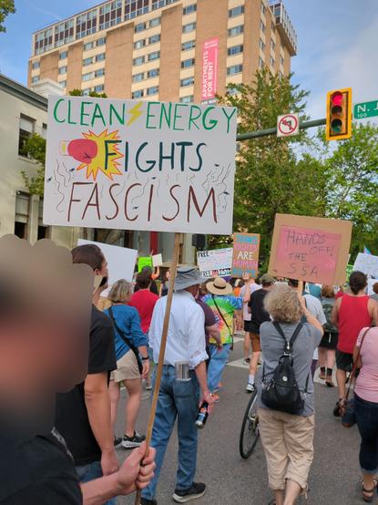 A man standing in a crowd of people protesting in the street. His sign says "clean energy fights fascism". His face is blurred.