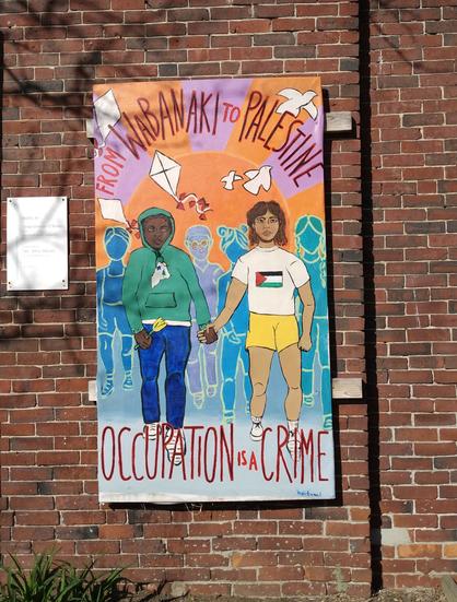 A painting mounted onto the side of a brick building.

The painting is of two people holding hands, with others behind them. There are doves and flags above them. The text reads:
"From Wabanaki to Palestine
Occupation is a Crime!"
