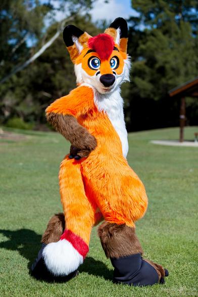 Colto Fox (orange, white, brown and red fox fursuiter) standing in full fursuit facing to to the right but looking directly at the viewer. He's standing in a park with his right arm on his hip. He's standing on grass and there are trees in the distant background.