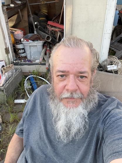 A bearded man in a gray shirt sits in a chair, looking directly at the camera. Behind him is a cluttered shed filled with various tools and materials, including buckets and pipes. Tall grass is visible in the foreground.