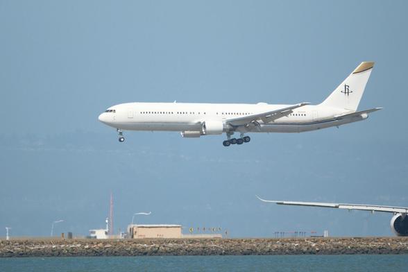 A white 767-300 with landing gear extended approaches a runway. The tail features a simple logo with the Houston Rockets initials, and a portion of another airplane's wing and engine is visible. That A330-900 aircraft has swooped up winglets. The background is a hazy sky with two thin white streamers on polls blowing toward the right.