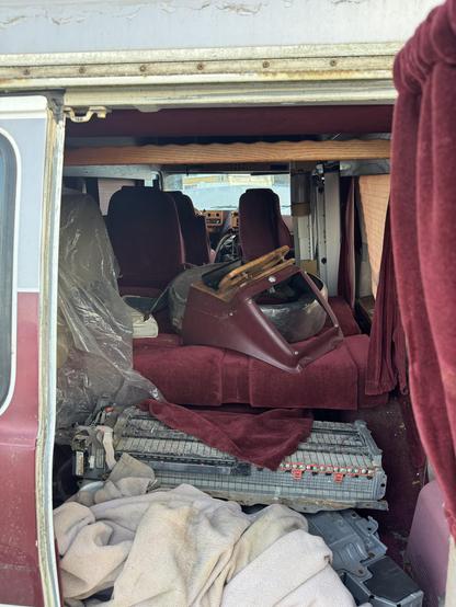 The image shows the interior of an old van with maroon upholstery. It features several seats, some covered in plastic, and a disassembled center console. Blankets and other items are scattered on the floor, suggesting disarray.