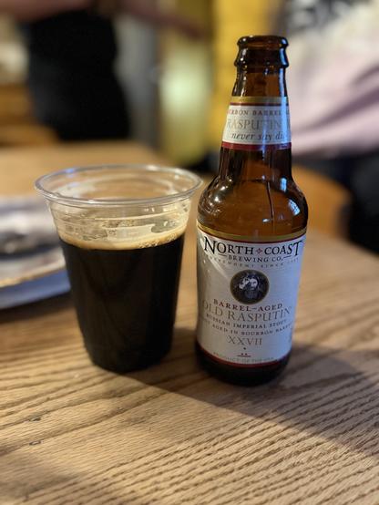 A plastic cup full of dark beer next to a bottle with a whites label and Rasputin making an “L” gesture atop his head